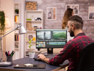 Back view of creative filmmaker working on a movie on laptop. Girlfriend in the background.